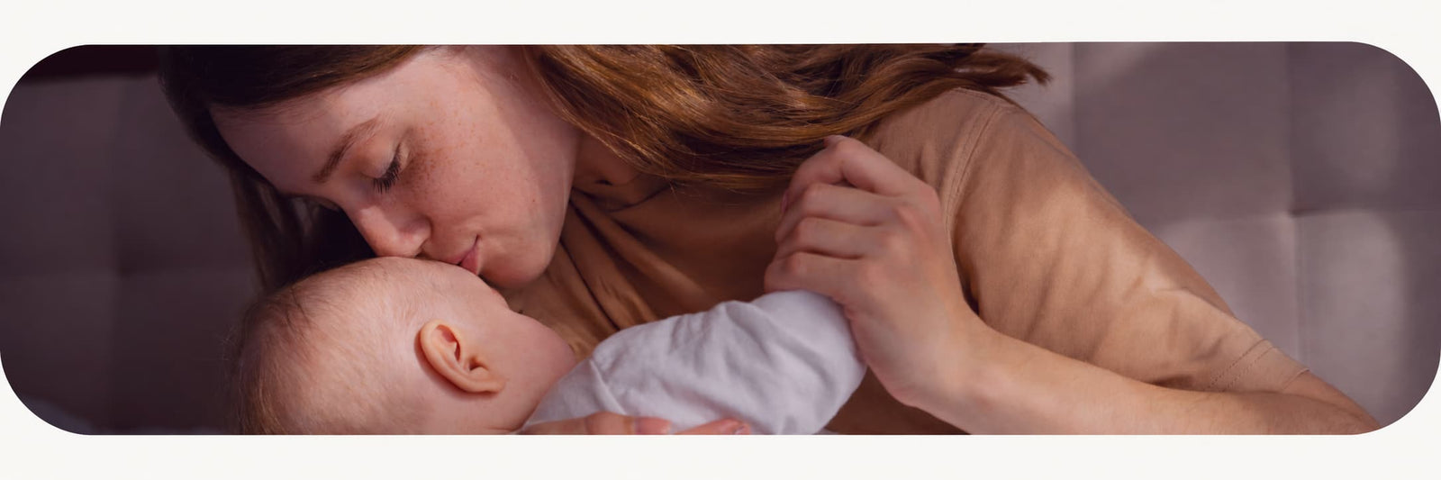 A woman breastfeeding a baby in a dimly lit, intimate setting. The woman has long light brown hair and is wearing a beige top, while the baby is dressed in a white outfit.