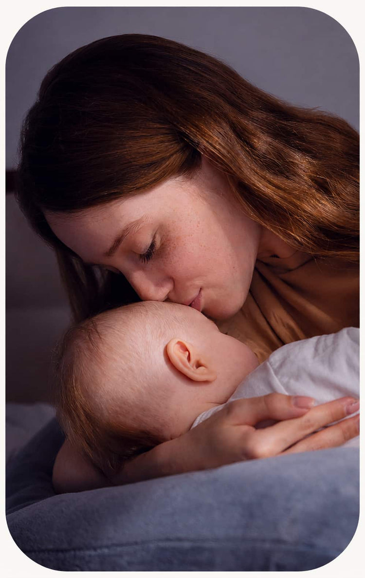 A woman breastfeeding a baby in a dimly lit, intimate setting. The woman has long light brown hair and is wearing a beige top, while the baby is dressed in a white outfit.