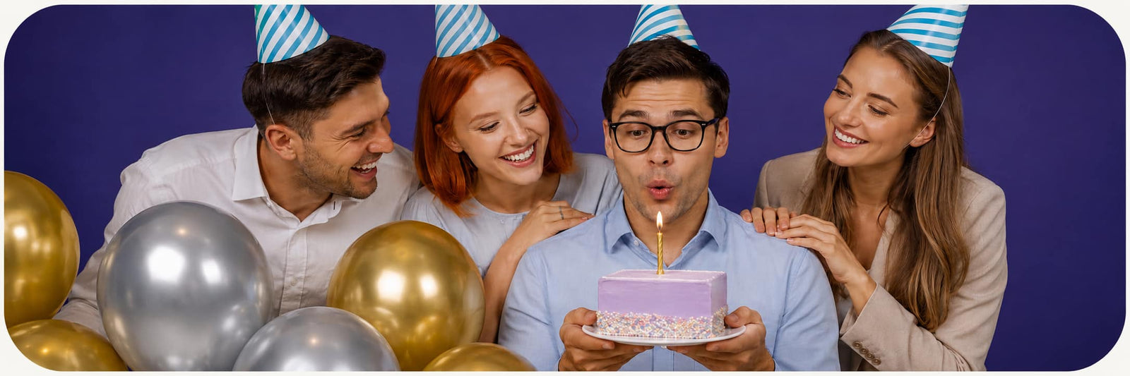 Four people wearing blue and white striped party hats gather around a man blowing out a small pink birthday cake with a candle. The group is surrounded by gold and silver balloons against a solid dark blue background.