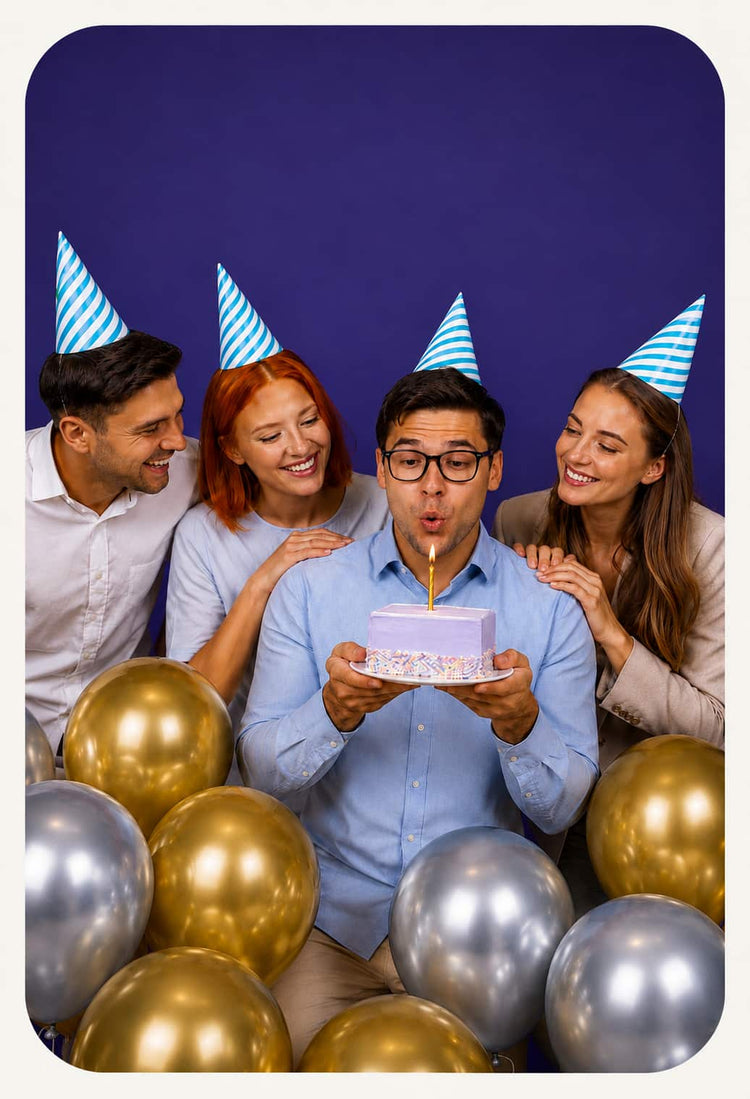 A group of four people wearing blue and white striped party hats celebrate with a small birthday cake and a single candle. The central person blows out the candle, surrounded by gold and silver balloons against a dark purple background.