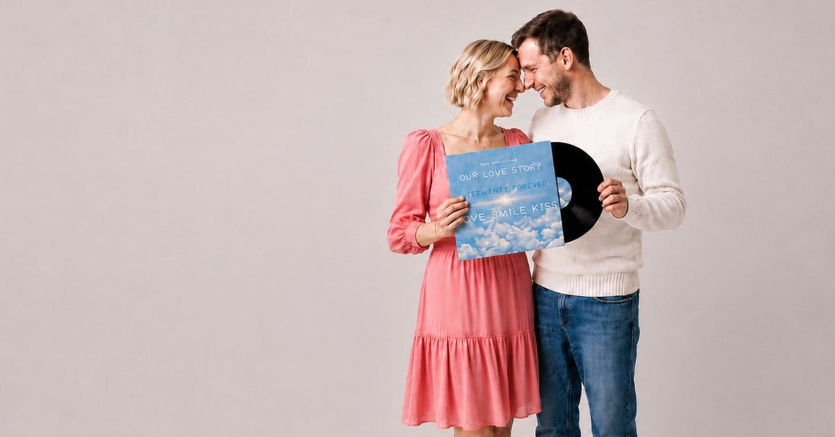 A man and a woman standing together against a light gray studio background, holding a blue vinyl record cover and part of a black vinyl record. The woman wears a pink dress, and the man wears a white sweater and blue jeans.