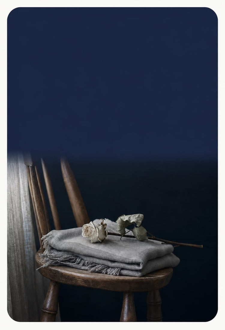 A folded gray linen throw blanket with fringe sits on a rustic wooden chair, topped with a dried white rose, against a dark navy blue background.